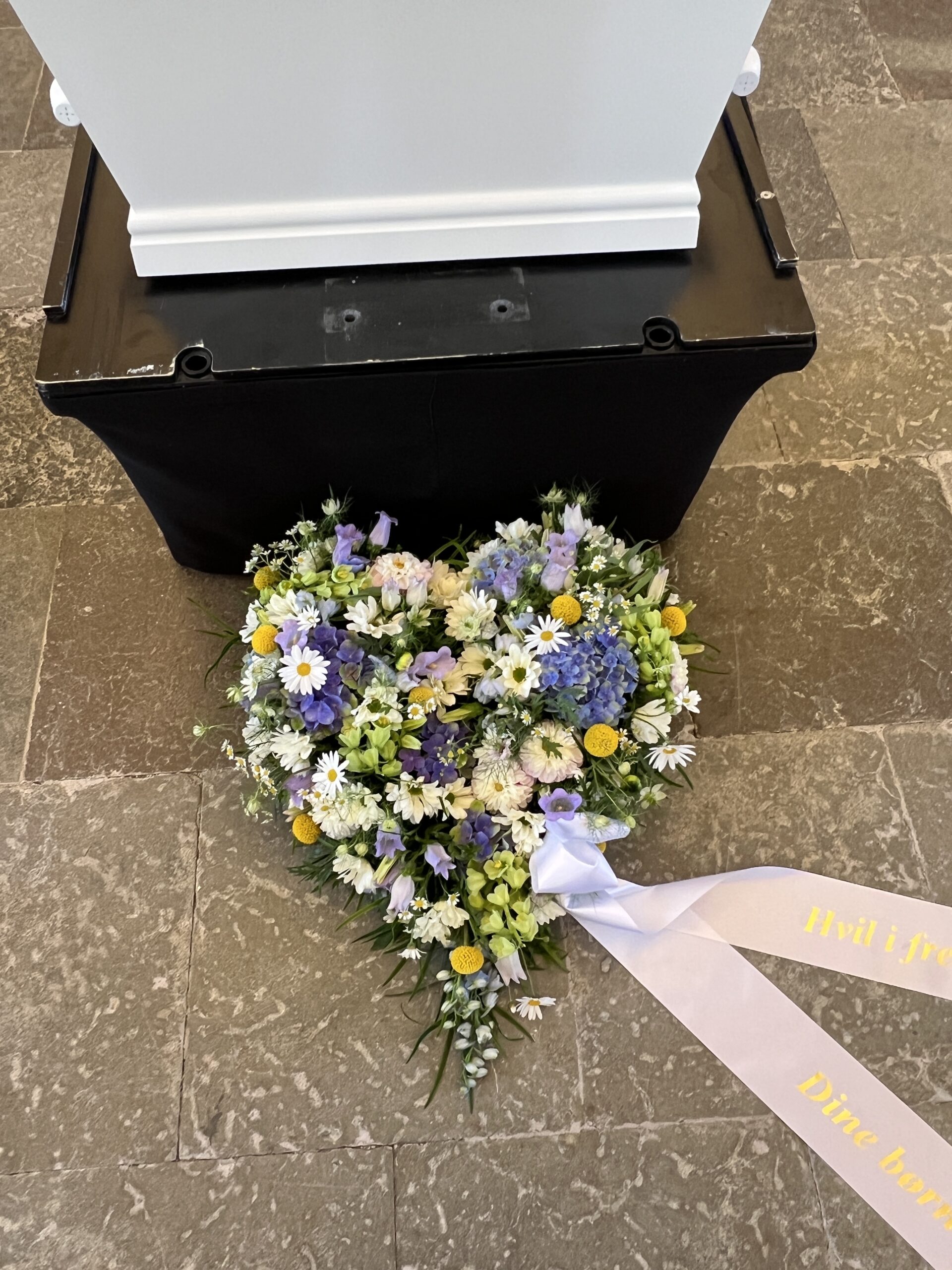 Blomsterhjerte i blå, hvide og gule nuancer lagt ved kisten
