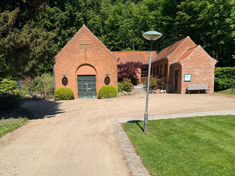 vedbaek_kapel | Kongevejens Bedemandsforretning Brick chapel with a cross above the arched green door, flanked by trimmed bushes on a sunny gravel path.