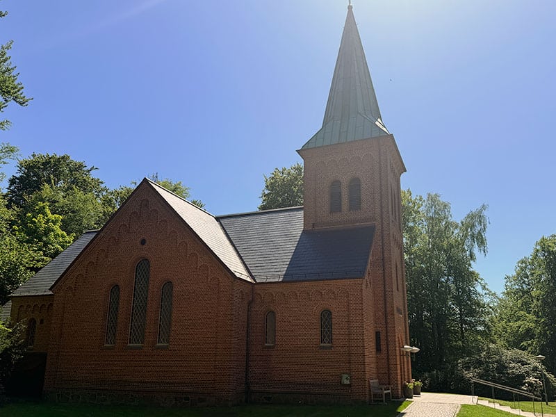 vedbaek_kirke_lille | Kongevejens Bedemandsforretning Brick church with a tall steeple under a blue sky, surrounded by trees on a sunny day