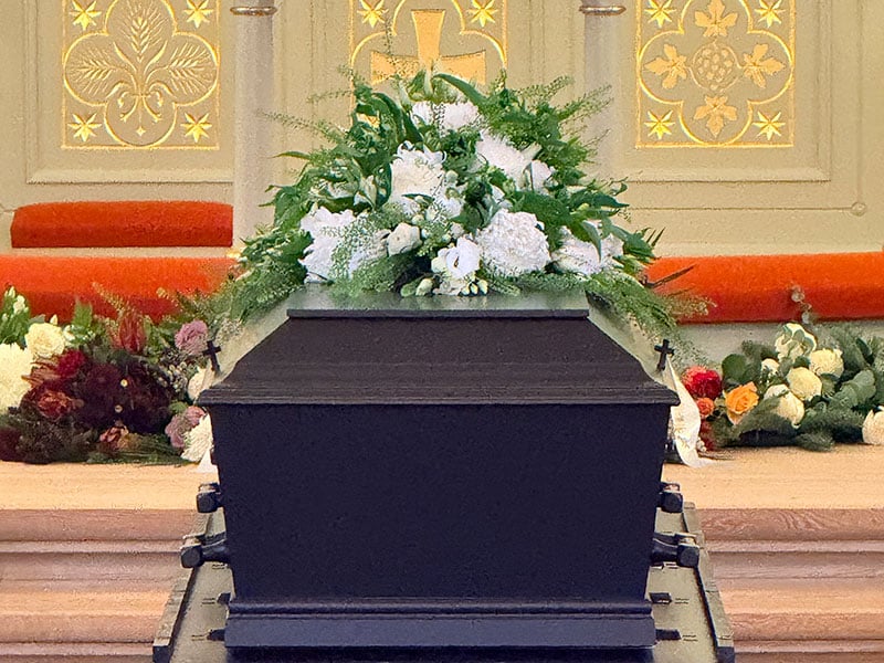 vedbaek_kiste | Kongevejens Bedemandsforretning Casket on a black bier with white flowers on top, placed at a church altar with floral arrangements in the background