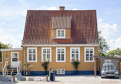 Yellow cottage with a red-tiled roof, white-framed windows, flower pots, gravel yard, and a car parked on the right.
